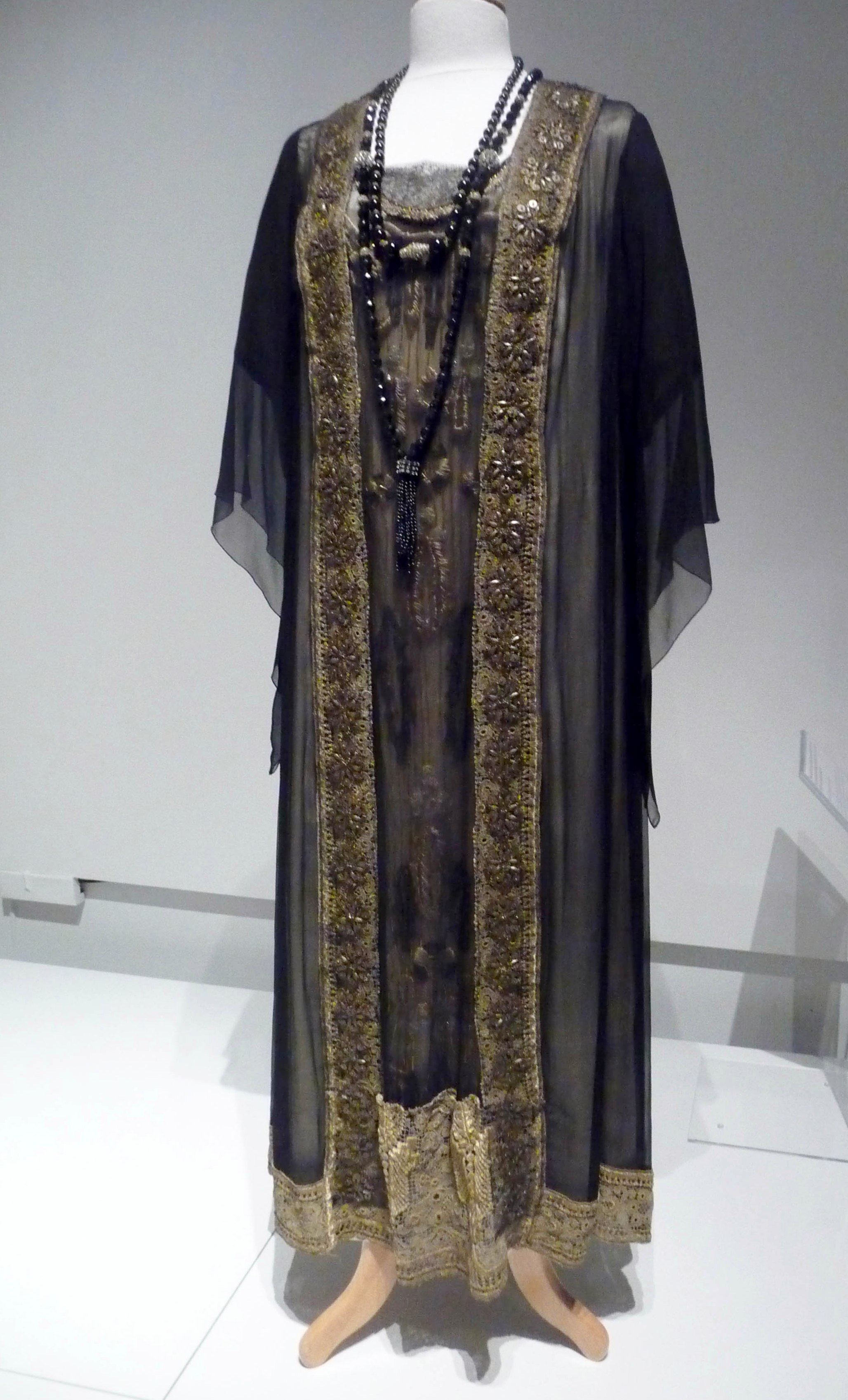 TUNIC-STYLE EVENING DRESS, cream silk under-dress with front panel embroidered in silk flock. Over-dress of black chiffon with gold metallic trim, made by Cosprop, 2012. Worn by Shirley MacLaine as Marthe Levinson in Downton Abbey.